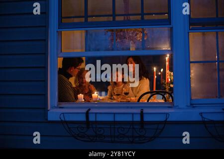 Famille profitant d'un dîner aux chandelles à l'intérieur d'une maison confortable vue de l'extérieur de la fenêtre bleue illuminée au crépuscule. Banque D'Images