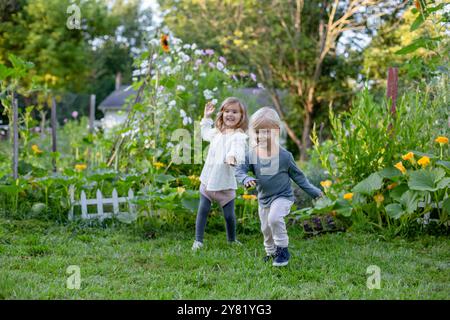 Deux enfants heureux courant main dans la main à travers un jardin luxuriant plein de fleurs et de plantes. Banque D'Images