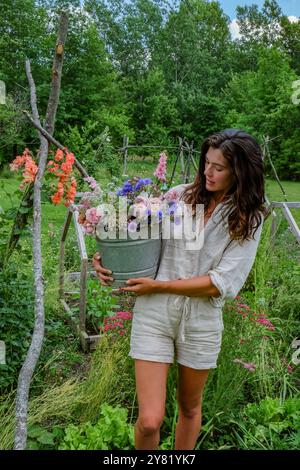 Femme tenant un seau plein de fleurs colorées dans un jardin luxuriant. Banque D'Images