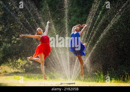 Deux femmes dans des robes colorées dansant joyeusement parmi les gouttelettes d'eau d'arrosage dans un cadre extérieur ensoleillé. Banque D'Images