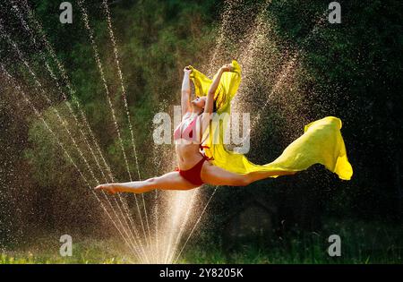 Femme en plein air effectuant un saut fendu avec un tissu jaune sur fond de saupoudrages d'eau ensoleillés. Banque D'Images