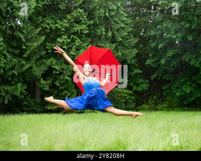 Femme joyeuse en robe bleue danse sur une pelouse verte avec un parapluie rouge. Banque D'Images