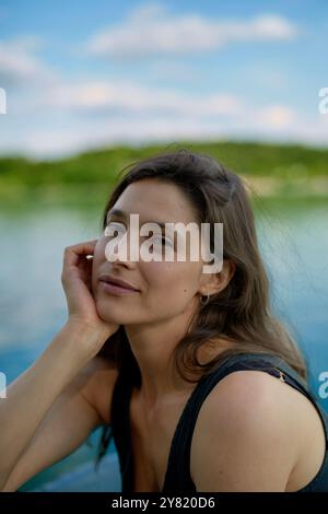 Femme souriante posant près d'un lac calme avec une expression sereine et sa main reposant sur sa joue. Banque D'Images