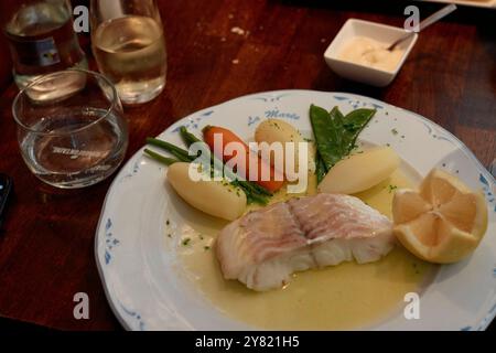 Un repas de poisson poché avec citron, pommes de terre bouillies, carottes et épinards servi sur une assiette blanche avec un verre d'eau et de vin blanc sur une table en bois. Banque D'Images