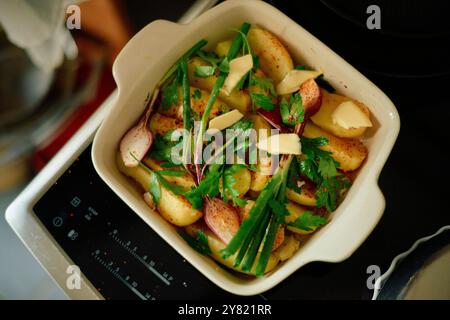 Tranches de pommes de terre cuites au four garnies de persil frais et de flocons de parmesan dans un plat blanc sur une cuisinière à induction. Banque D'Images