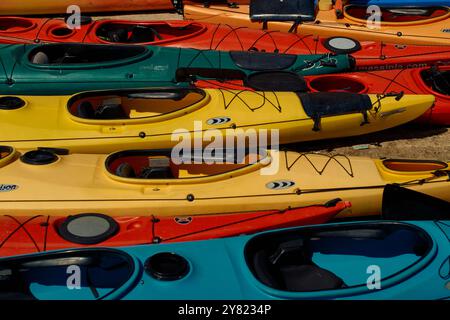 Kayaks colorés empilés sur une plage de sable sous la lumière du soleil, présentant une variété de designs et de tailles. Banque D'Images