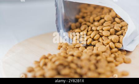 Gros plan d'un sac d'aliments pour animaux renversé, renversant son contenu sur une surface en bois. Les croquettes sont petites, rondes et brunes, créant une dispersion Banque D'Images