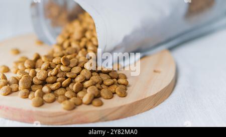 Un sac de nourriture pour animaux de compagnie renversé, renversant des croquettes sur une planche à découper en bois. Les croquettes sont petites, rondes et de couleur brun doré, avec un flou b Banque D'Images