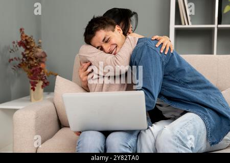 Jeune couple diversifié excité embrassant tout en travaillant avec un ordinateur portable. Partenaires romantiques féminins et masculins travaillant en ligne et célébrant le succès sur un canapé. Banque D'Images