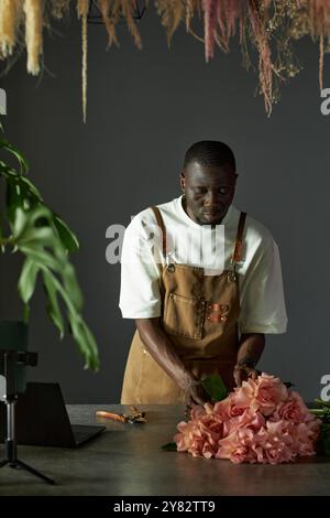 Portrait vertical du jeune homme afro-américain en tant que fleuriste masculin créant bouquet de roses et processus de tournage avec smartphone dans un cadre élégant Banque D'Images