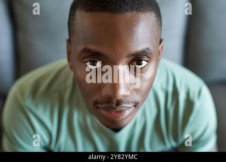 Gros plan portrait d'un jeune homme africain avec une expression sérieuse assis seul sur un canapé dans son salon à la maison Banque D'Images