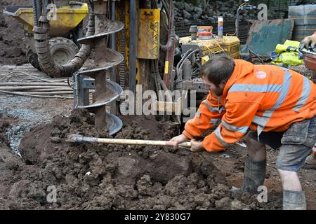 Homme déblayant manuellement les déblais de la machine de forage de pieux de tarière perçant le trou dans le sol argileux sur le chantier de construction domestique Banque D'Images