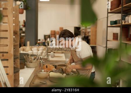 Jeune céramiste masculin portant un tablier façonnant une argile dans un bol sur une roue de poterie dans un atelier de céramique Banque D'Images