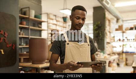 Jeune ouvrier de studio de céramique africain debout dans un grand atelier de fabrication de poterie et utilisant une tablette numérique Banque D'Images