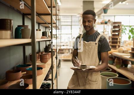 Jeune ouvrier de studio de poterie africaine avec une planche à pince faisant l'inventaire des bols assis sur les étagères dans un atelier Banque D'Images