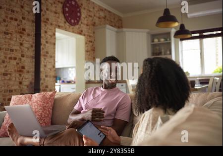 Rire jeune couple multiracial utilisant un ordinateur portable et une tablette numérique tout en se relaxant ensemble sur leur canapé de salon Banque D'Images