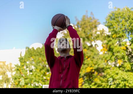 Beau joueur de basket-ball tirant une balle à travers le cerceau tout en jouant sur un terrain de basket-ball à l'extérieur Banque D'Images