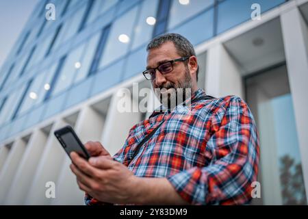 Portrait de bel homme d'affaires avec lunettes de soleil et chemise à carreaux marchant dans la rue et tapant des messages sur son téléphone portable. Mature Casually Dresse Banque D'Images