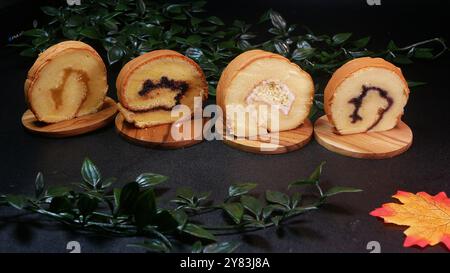 Quatre tranches molles de gâteau roulé avec différentes garnitures, allant de la confiture de fruits à la crème de noix, servies sur des planches de bois avec un fond de feuille verte Banque D'Images