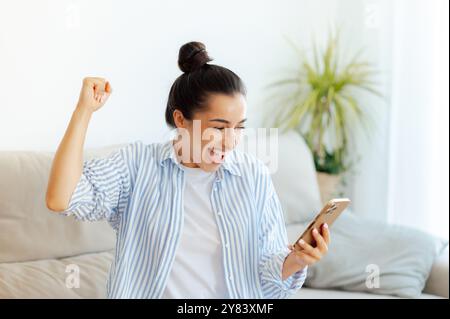 Joyeuse jeune femme caucasienne heureuse en tenue décontractée, tient un smartphone lisant de bonnes nouvelles, célébrant la victoire, gagnant le jeu, se sentir heureuse assis sur le canapé à la maison, faisant des gestes avec le poing, souriant Banque D'Images