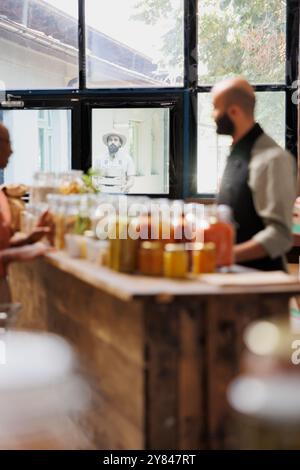 La cliente interagit avec un ouvrier de magasin masculin, entre eux se trouve une table avec des pots en verre remplis de plusieurs produits bio-alimentaires. En arrière-plan, homme avec un chapeau transportant des boîtes à l'extérieur du magasin. Banque D'Images