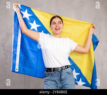 Heureuse jeune femme avec drapeau de Bosnie-Herzégovine sur fond de mur gris Banque D'Images