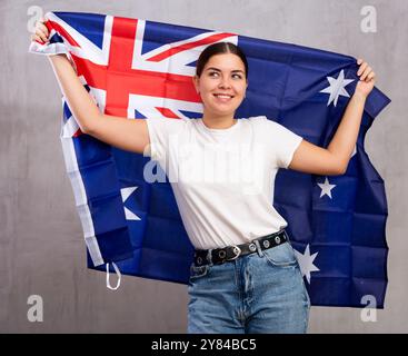 Joyeuse jeune femme agitant le drapeau national de l'Australie tout en se tenant debout contre le mur gris à l'intérieur Banque D'Images