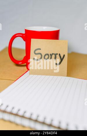Phrase de pardon écrite sur un carton recyclé avec une tasse de café rouge sur le fond d'un cahier. Banque D'Images