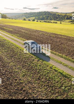 Une voiture roule sur une piste de terre entre de grands champs sous un ciel avec nuages et soleil du soir, partage de voiture, VW ID5, Calw, Forêt Noire, Allemagne, Europe Banque D'Images