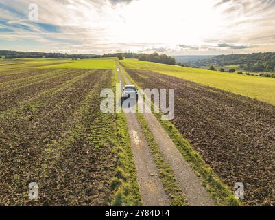 Voiture noire conduisant sur une longue voie de campagne droite à travers des champs panoramiques sous un ciel nuageux, partage de voiture, VW ID5, Calw, Forêt Noire, Allemagne, Europe Banque D'Images
