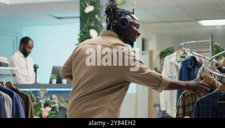Homme afro-américain dansant dans la boutique de mode fantaisie tout en recherchant des vêtements habillés élégants. Client heureux écoutant de la musique dans des écouteurs tout en achetant des vêtements de créateurs dans une salle d'exposition haut de gamme Banque D'Images