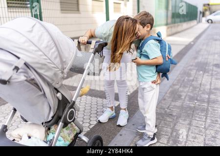 Mère embrassant son fils allant au premier jour à l'école debout au milieu de la rue et tenant une voiture d'enfant Banque D'Images