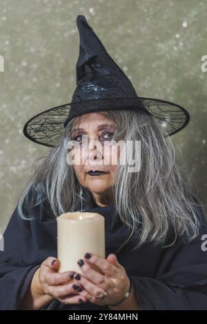 Gros plan d'une vieille sorcière aux cheveux blancs vêtue de noir avec un chapeau et une grande bougie blanche dans ses mains Banque D'Images