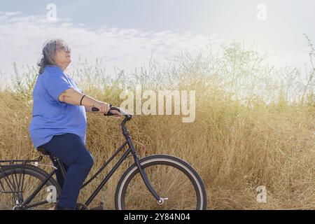 Une femme âgée aux cheveux blancs, obèse, vue de profil à vélo le long d'un sentier ensoleillé et envahi par la végétation Banque D'Images
