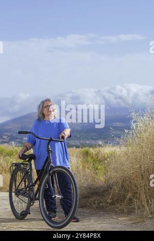 Femme mature souriante en surpoids chevauchant un vélo sur une route de campagne faisant de l'exercice, en arrière-plan un paysage montagneux avec des nuages. concept health c Banque D'Images