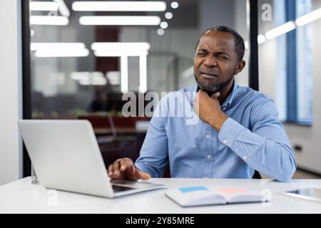 Homme en chemise bleue assis au bureau à l'aide d'un ordinateur portable, affichant un inconfort de gorge clair dans un bureau moderne. Ordinateur portable et portable sur table créent un cadre professionnel, transmettant le stress et les problèmes de santé Banque D'Images