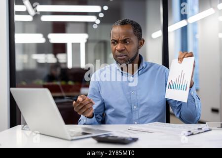 Homme d'affaires étudie attentivement le rapport financier avec graphique à barres sur papier pendant la réunion en ligne. Professionnel portant chemise bleue dans un environnement de bureau moderne, expression montrant la concentration. Banque D'Images