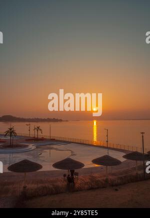 Un coucher de soleil doré serein jette des reflets sur la mer calme, avec des palmiers et des parasols créant une atmosphère tranquille pour la détente. Banque D'Images