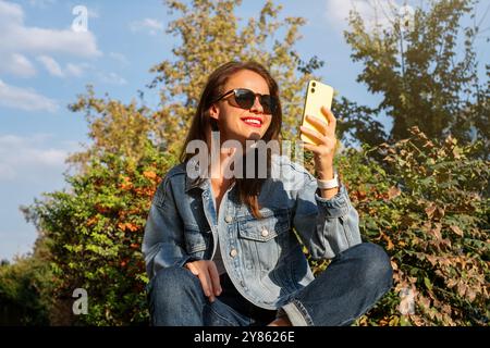 Une femme portant une veste en denim et des lunettes de soleil est assise sur un banc de parc et utilise un smartphone. Banque D'Images