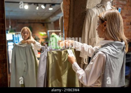 Femme réfléchie magasinant pour de nouveaux vêtements dans le magasin, tient cintre de pantalons en coton vérifiant les étiquettes de prix Banque D'Images