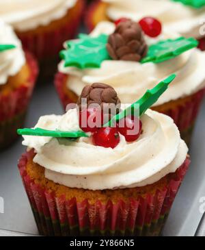 Des cupcakes de Noël avec glaçage blanc, décorés de baies rouges, de pommes de pin au chocolat et de feuilles de houx attendent sur un plateau pour la partie de noël Banque D'Images