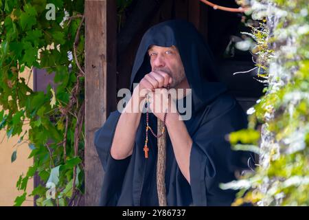 Un moine dans une robe noire tenant un chapelet de prière sur un fond d'une vieille maison et de verdure. Banque D'Images