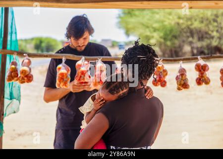 Vendeur de rue africain de village, mère tenant sa fille dans ses bras tous les deux vêtus de cheveux tressés, jeune homme caucasien achetant des tomates Banque D'Images