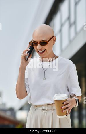 Vêtue de vêtements élégants, une femme chauve sourit largement alors qu'elle discute au téléphone, tenant un café. Banque D'Images