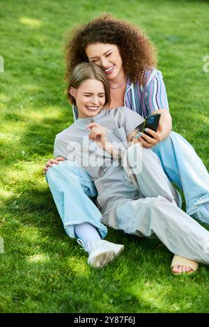 Deux amis partagent un moment joyeux sur l'herbe, riant et regardant un téléphone ensemble. Banque D'Images