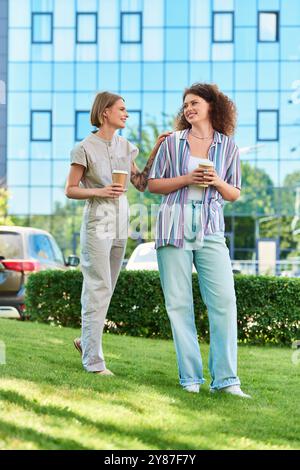 Deux amis partagent le rire et le café alors qu'ils marchent dans un parc urbain verdoyant par une journée ensoleillée. Banque D'Images