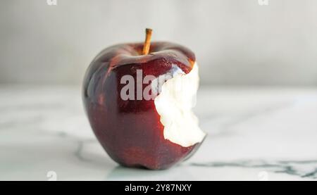 Pomme brillante rouge foncé mordue sur fond de surface en marbre blanc, image minimale et conceptuelle symbolisant la tentation, fruit sain avec une peau brillante, a Banque D'Images