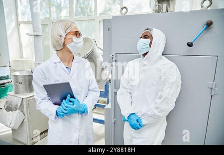 Équipe diversifiée de deux travailleurs professionnels de l'industrie pharmaceutique en uniforme souriant tout en communiquant sur le plancher de production à l'usine de fabrication Banque D'Images