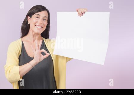 Femme souriante tient un signe blanc avec une main et avec l'autre main elle fait le symbole OK. Image pour annoncer des offres et des actualités commerciales. Banque D'Images