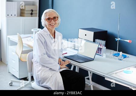 Portrait moyen de chimiste de laboratoire féminine souriante dans une résille médicale assise à la table de travail avec ordinateur portable dans le laboratoire du service de recherche scientifique, espace de copie Banque D'Images